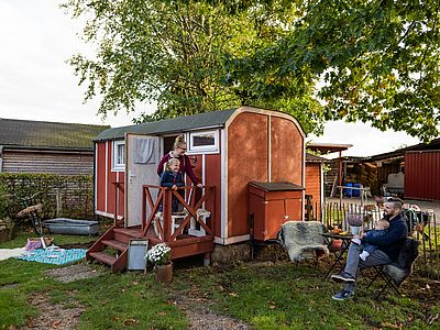 Eine Familie genießt ihren Bauernhofurlaub vor einem roten Ferienwagen. Eine Frau steht mit einem Kind auf der kleinen Veranda, während ein Mann mit einem Kleinkind draußen sitzt.