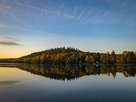 Salemer See bei tief stehender Sonne mit ruhiger Wasseroberfläche. Der herbstlich gefärbte Wald am Ufer spiegelt sich deutlich im See.