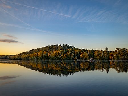 Salemer See bei tief stehender Sonne mit ruhiger Wasseroberfläche. Der herbstlich gefärbte Wald am Ufer spiegelt sich deutlich im See.
