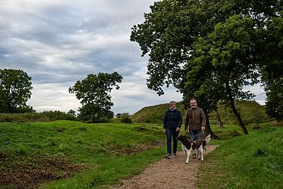 Zwei Erwachsene gehen gemeinsam mit einem Hund an der Leine über einen schmalen Weg durch eine grüne Landschaft. Rundherum wachsen Wiesen und mehrere große Bäume.
