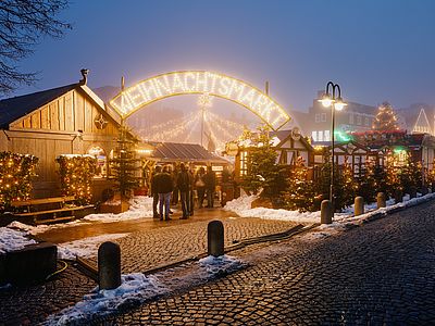 Festlich beleuchteter Weihnachtsmarkt in der Dämmerung mit Holzhütten, geschmückten Tannenbäumen und einem leuchtenden Eingangsbogen. Besucher schlendern zwischen den Ständen, während Schnee die Wege säumt.