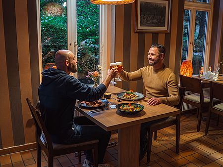 Zwei Personen sitzen sich in einem gemütlich eingerichteten Restaurant an einem Holztisch gegenüber und stoßen mit Biergläsern an. Auf dem Tisch stehen zwei Hauptgerichte und ein Salat sowie eine kleine Blumenvase. Im Hintergrund sorgen große Fenster mit Blick ins Grüne und warme Tischlampen für eine ruhige, einladende Atmosphäre.