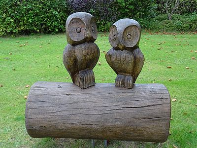 Zwei aus Holz geschnitzte Eulen sitzen nebeneinander auf einem waagerechten Baumstamm vor einer grünen Wiese.