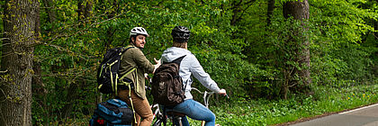 Zwei Radfahrende fahren mit Rucksack und Helm entlang eines Waldes. 