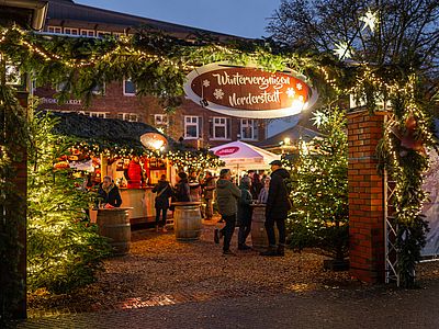 Festlich geschmückter Eingang zum Weihnachtsmarkt „Wintervergnügen Norderstedt“ mit beleuchteten Tannenbäumen und Lichterketten. Besucher stehen an den Weihnachtsmarktständen und unterhalten sich an Stehtischen.