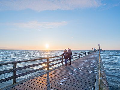 Zwei Personen stehen eng beieinander auf einer langen Holzseebrücke und blicken bei tief stehender Sonne über das Meer. Auf den Holzplanken liegen vereinzelte Schneereste.
