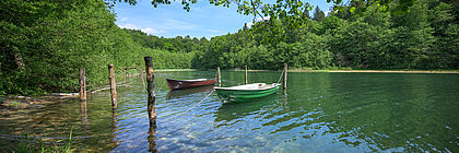 Ein klarer See ist von Wald umgeben. Auf dem See sind ein rotes und ein grünes Boot an Baumstämmen befestigt. - zum Artikel 'Wunderbar Wandern im Naturschutzgebiet Salemer Moor '