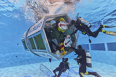Sieben Menschen trainieren auf dem GreenTEC Campus Unterwasser in einem Wasserbecken Extremsituationen, einige befinden sich in einer Art Rettungskapsel, eine Hubschrauberkapsel.