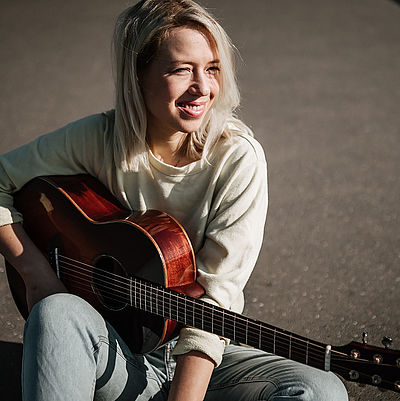 Eine junge Frau aus Föhr mit längeren blonden Haaren sitzt lachend draußen auf dem Boden, blickt in die Ferne und hält eine Gitarre in der Hand.