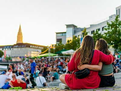 Zwei Frauen sitzen Arm in Arm am Kieler Bootshafen und blicken auf die Veranstaltung. Im Hintergrund ist der Kieler Rathausturm zu sehen.