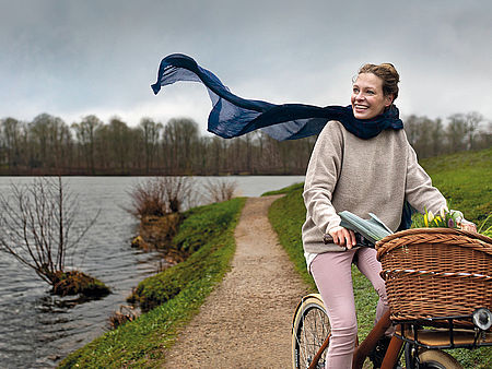 Eine Frau fährt auf dem Fahrrad einen Weg am Ufer eines Gewässers entlang. Ihr Schal weht im Wind.