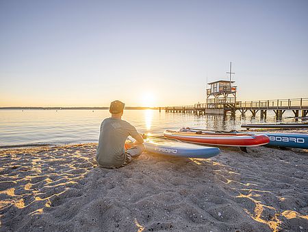 Eine Person sitzt mit dem Rücken zur Kamera am Sandstrand und blickt auf ruhiges Wasser bei Sonnenuntergang. Neben ihr liegen mehrere Stand-up-Paddleboards im Sand. Rechts führt ein langer Holzsteg zu einem kleinen Rettungsturm über dem Wasser.
