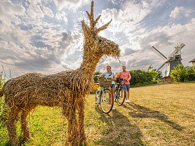 Zwei Personen gehen mit ihren Fahrrädern an einer Hirsch-Kornfigur vorbei, im Hintergrund eine alte Mühle.