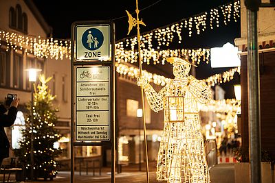 Leuchtende Figur eines Nachtwächters mit Laterne und Hellebarde in einer weihnachtlich geschmückten Fußgängerzone. Im Hintergrund erhellen Lichterketten und ein geschmückter Tannenbaum die Straße.