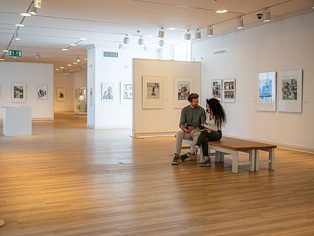 Zwei Personen sitzen auf einer Bank in einem hellen Ausstellungsraum und betrachten gemeinsam einen Museumsführer. An den Wänden hängen gerahmte Kunstwerke, weitere Ausstellungsbereiche erstrecken sich im Hintergrund.