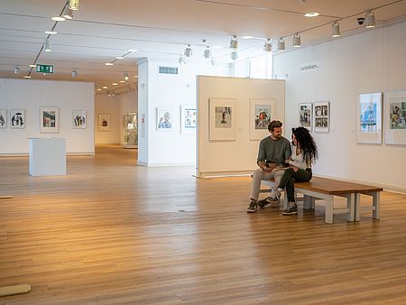 Zwei Personen sitzen auf einer Bank in einem hellen Ausstellungsraum und betrachten gemeinsam einen Museumsführer. An den Wänden hängen gerahmte Kunstwerke, weitere Ausstellungsbereiche erstrecken sich im Hintergrund.