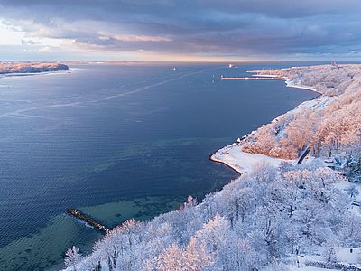 Eine verschneite Küstenlandschaft aus der Vogelperspektive. Schnee bedeckt die bewaldeten Ufer, während sich das ruhige, tiefblaue Meer unter einem wolkenverhangenen Himmel bis zum Horizont erstreckt.
