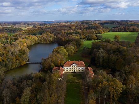 Luftaufnahme eines Herrenhauses mit rotem Dach, umgeben von herbstlich gefärbten Wäldern, grünen Feldern und einem See mit kleiner Brücke.