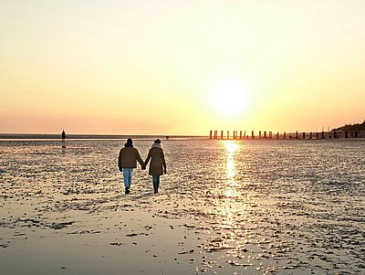Zwei Personen gehen Hand in Hand über das Watt bei Sonnenuntergang. Die tief stehende Sonne spiegelt sich auf der feuchten Oberfläche des Wattenmeers und taucht die Landschaft in warmes, goldenes Licht.