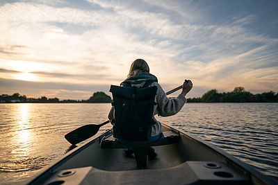 ine Person sitzt in einem Kanu auf einem ruhigen See und paddelt. Sie trägt einen Rucksack und warme Kleidung. Vor ihr liegt die Wasserfläche, am Horizont sind Bäume zu sehen.