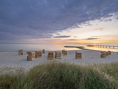 Blick über einen breiten Sandstrand an der Ostsee bei Sonnenuntergang. Mehrere Strandkörbe stehen verteilt auf dem hellen Sand. Im Vordergrund wachsen Dünengräser, rechts führt ein langer Steg ins ruhige Meer.
