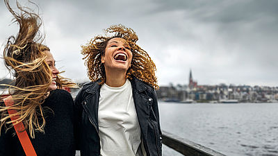Zwei lachende Frauen gehen auf einem Steg entlang, im Hintergrund ist eine Stadt zu sehen. Die Haare der beiden Frauen wehen im Wind. 