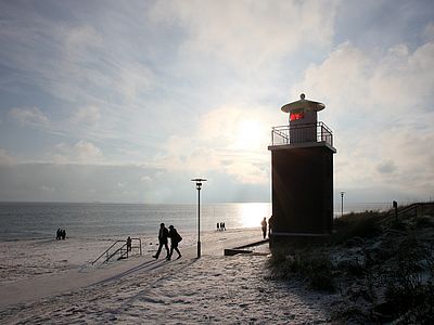 Ein kleiner Leuchtturm steht am verschneiten Strand, im Hintergrund glitzert die tiefstehende Wintersonne auf der Wasseroberfläche. Mehrere Menschen spazieren am Ufer entlang.