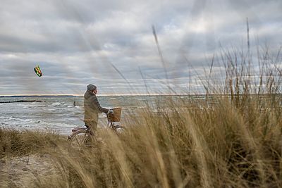 Eine Person mit Winterjacke, Handschuhen und Mütze steht am Strand neben einem Fahrrad mit Korb. Im Vordergrund wachsen hohe Gräser, im Hintergrund sind die raue See, Wellen und ein Kitesurfer mit buntem Kite zu sehen.