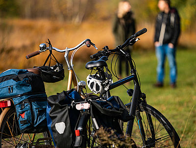 Zwei Fahrräder mit Gepäcktaschen und Fahrradhelmen am Lenker. Im Hintergrund stehen zwei Personen.