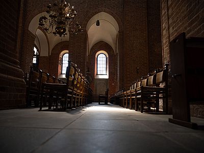 Blick durch den Mittelgang des Ratzeburger Doms auf Stuhlreihen, Backsteinbögen, hohe Fenster und einen verzierten Kronleuchter.