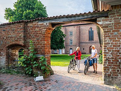 Zwei Erwachsene schieben ihre Fahrräder durch einen Torbogen aus Backstein auf den Innenhof eines historischen Klosters.