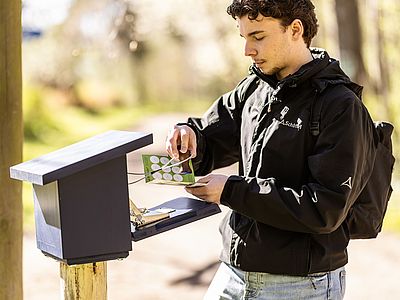 Eine Person steht im Freien an einer hölzernen Station entlang eines Weges und stempelt eine grüne Karte. Sie trägt eine schwarze Outdoorjacke und einen Rucksack. Die Stempelstation ist auf einem Holzpfosten montiert.