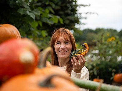 Eine Frau hält lächelnd einen kleinen gelb-grünen Zierkürbis in der Hand, umgeben von orangefarbenen Kürbissen.