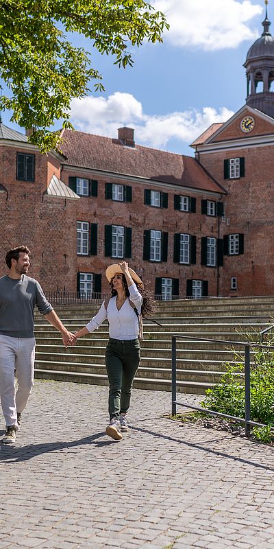 Ein Paar geht Hand in Hand über einen gepflasterten Weg vor dem Eutiner Schloss. Im Hintergrund ist der rote Backsteinbau mit seinem markanten Turm zu sehen.