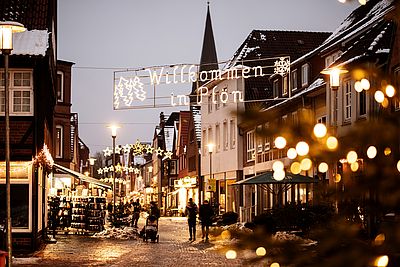 Weihnachtlich beleuchtete Fußgängerzone in Plön mit dem leuchtenden Schriftzug „Willkommen in Plön“ und Sternen über der Straße. Passanten schlendern zwischen den Geschäften, während Schnee die Straßenränder und Dächer bedeckt.
