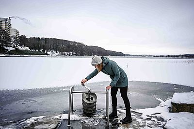 Eine Frau in Winterkleidung steht am Großen Segeberger See und dreht eine spiralförmige Metallkonstruktion in den gefrorenen See.