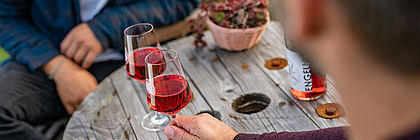 Zwei gefüllte Gläser mit Rotwein und eine Rotweinflasche stehen auf einem Holztisch. Eine männliche Hand hält ein Glas fest. - zum Artikel 'Willkommen in der nördlichsten Weinregion Deutschlands'