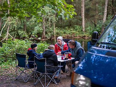 Eine Familie sitzt beim Camping an einem Tisch im Wald und spielt gemeinsam. Auf dem Tisch stehen Getränke, daneben ist ein blauer Campingbus geparkt.
