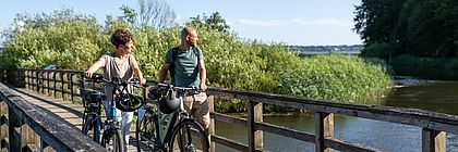 Eine Frau und ein Mann gehen mit ihren Fahrrädern über eine Holzbrücke und blicken über das von Schilf gesäumte Wasser. Im Hintergrund erstreckt sich eine grüne Uferlandschaft unter blauem Himmel. - zum Artikel 'Unterwegs auf zwei Rädern zwischen Nordsee und Ostsee: Fahrradtour auf dem Wikinger-Friesen-Weg '