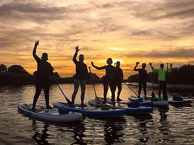 Sechs Personen stehen auf Stand-up-Paddleboards auf einem ruhigen Gewässer und heben teilweise die Hand zum Winken. Sie sind als dunkle Silhouetten vor einem orange-goldenen Sonnenuntergang zu sehen.