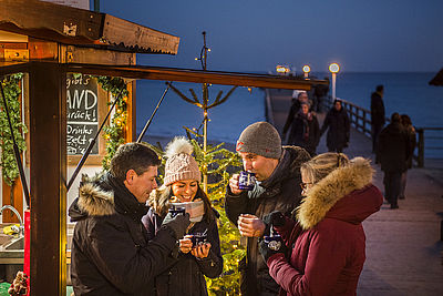Zwei Männer und zwei Frauen stehen an einer Punschbude und trinken ein heißes Getränk. Im Hintergrund befindet sich eine Seebrücke.
