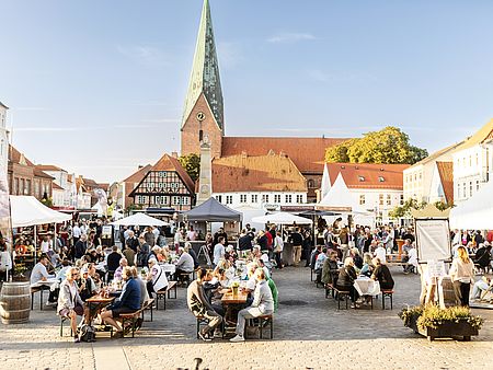 Belebter Marktplatz bei sonnigem Wetter: Zahlreiche Menschen sitzen an langen Holzbänken oder gehen zwischen weißen Marktständen umher. Im Hintergrund steht eine große Backsteinkirche mit hohem, spitzem Turm. Umgeben wird der Platz von historischen Gebäuden mit roten Ziegeldächern.