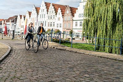 Eine Frau und ein Mann fahren mit Fahrrädern über eine gepflasterte Straße in Friedrichstadt. Im Hintergrund stehen historische Giebelhäuser; eine große Trauerweide säumt den Weg.