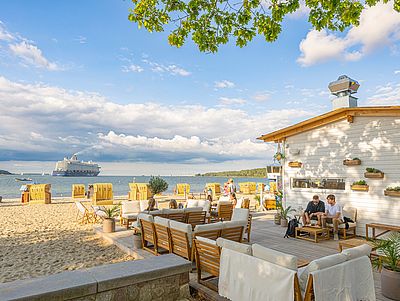 Strandterrasse eines Cafés mit Sitzgelegenheiten direkt am Sandstrand. Im Hintergrund stehen gelbe Strandkörbe am Ufer, auf der Ostsee fährt ein Kreuzfahrtschiff.
