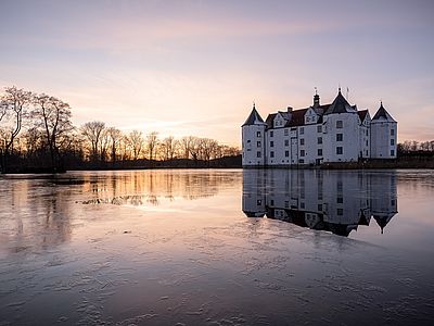 schreibe einen barrierefreien Alternativtext zu diesem Bild: Das weiße Wasserschloss Glücksburg mit seinen markanten Türmen spiegelt sich bei Sonnenuntergang im teilweise zugefrorenen Schlossteich.