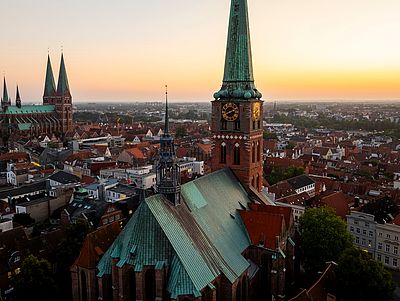 Luftaufnahme der historischen Altstadt von Lübeck bei Sonnenuntergang. Dächer, historische Gebäude und markante Türme prägen das Stadtbild unter einem warmen Abendhimmel.