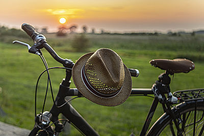 Ein Sommerhut hängt über dem Lenker eines Fahrrads. Im Hintergrund ist ein Sonnenaufgang.ein 