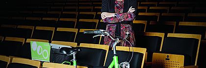 Illona Jarabek steht in einem Konzertsaal zwischen den Stühlen. Ein grünes Fahrrad steht in der Stuhlreihe vor ihr. - zum Artikel 'Nachhaltiges Dreamteam: Ilona Jarabek und die MUK Lübeck'