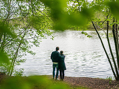Ein Mann und eine Frau stehen Arm in Arm an einem See und schauen auf das Wasser.