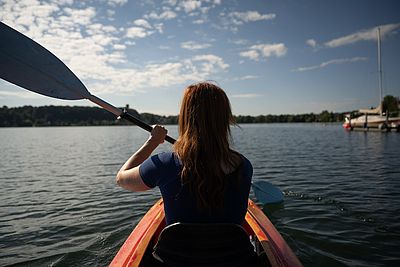 Eine Frau paddelt bei sonnigem Wetter mit einem roten Kajak über einen See. Am Ufer liegen mehrere Segelboote.
